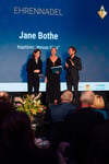 Das eloquente Moderatorenpaar der MV-Tourismustage, Anne Stadtfeld und Tobias Woitendorf, gratulieren der Kapitänin der Weissen Düne, Jane Bothe, zur Ehrennadel 2024 für besondere Verdienste.